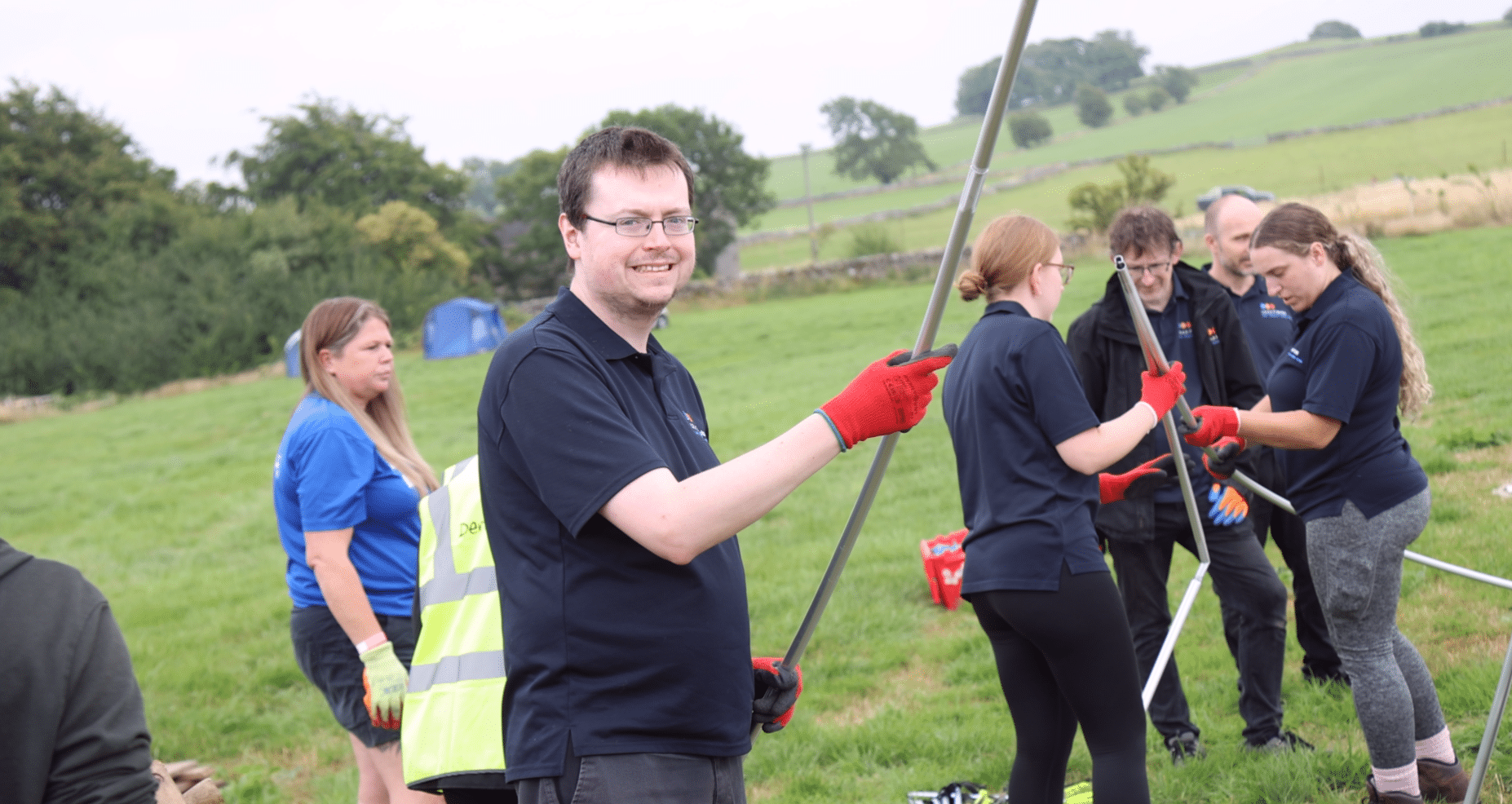 A man standing in a field smiling, wearing glasses and a blue L.E.A.D. IT work polo and red work gloves, holding a tent pole in both hands and smiling. A lady in the backgruond (left) with brown hair and wearing a blue tshirt. On the right in the background 4 L.E.A.D. IT staff members holding a longer tentpole bent in two, two women and two men.