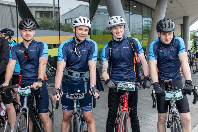 4 men on bikes front facing, with helments on and dark blue branded L.E.A.D. IT Services cycling jerseys,
