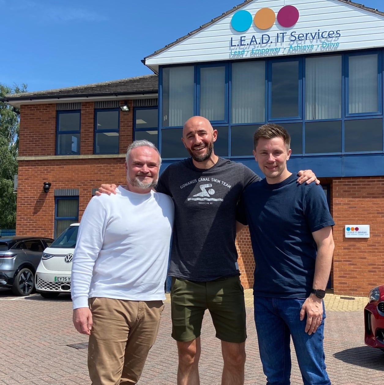 Rebuild East Midlands L.E.A.D. IT Services Partnership image with Chris Edwards (left) in a white long sleeve t-shirt and beige trousers, Paul McAnulty (center) in a dark grey short-sleeve t-shirt that reads 'GOWANUS CANAL SWIM TEAM' with a sign-like image depicting a person swimming in water and green shorts and Lee Jepson (right) in a dark blue short-sleeve t-shirt and dark blue jeans. All standing in front of the L.E.A.D. IT Services office